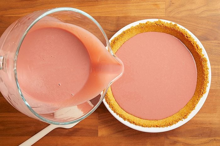 Overhead horizontal image of a pink-colored pie filling being poured from a glass measuring cup into a golden brown graham cracker pie crust set in a white pie plate. The smooth filling begins to spread within the crust, which rests on a wooden surface.