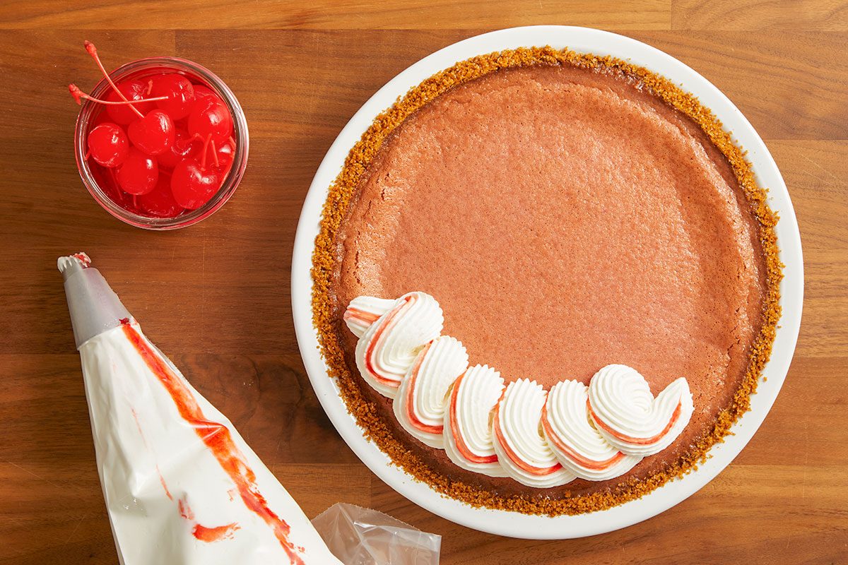 Overhead horizontal image of a chocolate pie with a graham cracker crust being decorated with piped whipped cream that has red streaks. A piping bag with a star tip and a glass jar of maraschino cherries are also visible on the wooden surface.