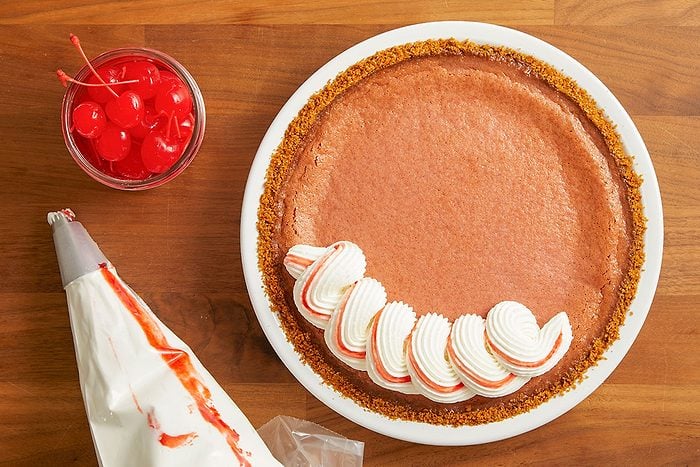 Overhead horizontal image of a chocolate pie with a graham cracker crust being decorated with piped whipped cream that has red streaks. A piping bag with a star tip and a glass jar of maraschino cherries are also visible on the wooden surface.
