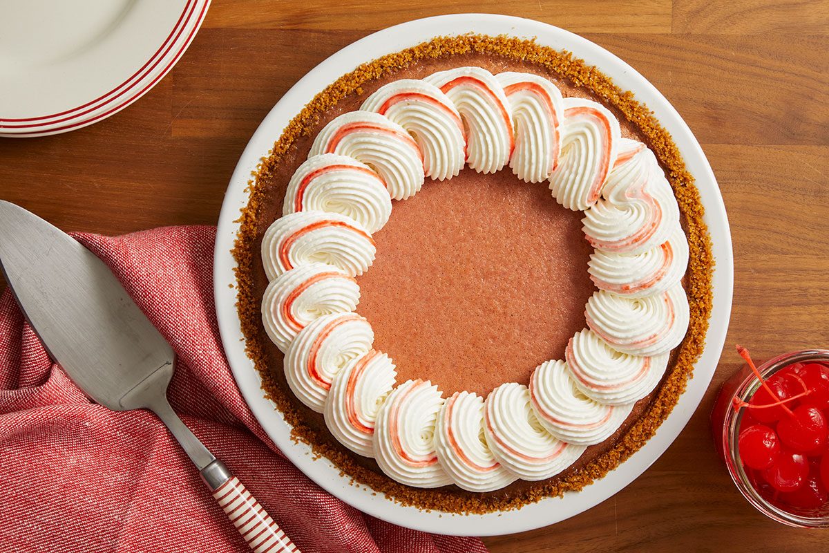 Overhead horizontal image of a chocolate or mocha pie with a graham cracker crumb crust, decorated with a circle of white whipped cream swirls, each featuring a reddish stripe. A pie server with a red and white striped handle, a red napkin, and a small jar of maraschino cherries are visible on a wooden surface.