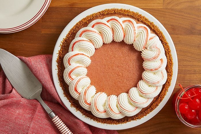 Overhead horizontal image of a chocolate or mocha pie with a graham cracker crumb crust, decorated with a circle of white whipped cream swirls, each featuring a reddish stripe. A pie server with a red and white striped handle, a red napkin, and a small jar of maraschino cherries are visible on a wooden surface.