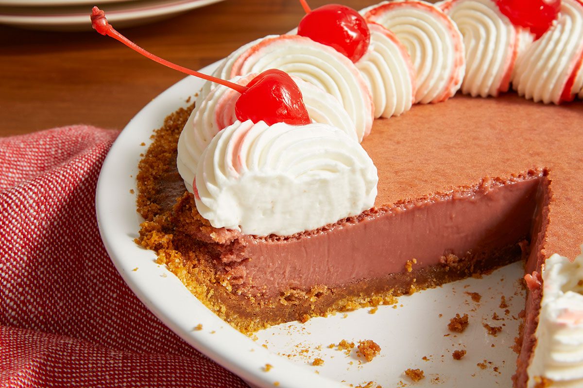 Eye Level horizontal image of a dessert pie with a slice removed, revealing its crumbly crust and reddish-brown filling. The pie is topped with swirls of white whipped cream, some with pink accents, and garnished with bright red maraschino cherries.