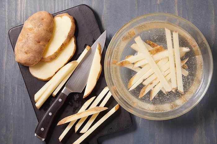 A cutting board with a whole potato, potato slices, a knife, and raw potato strips next to a bowl of water containing more potato strips, all on a wooden surface.