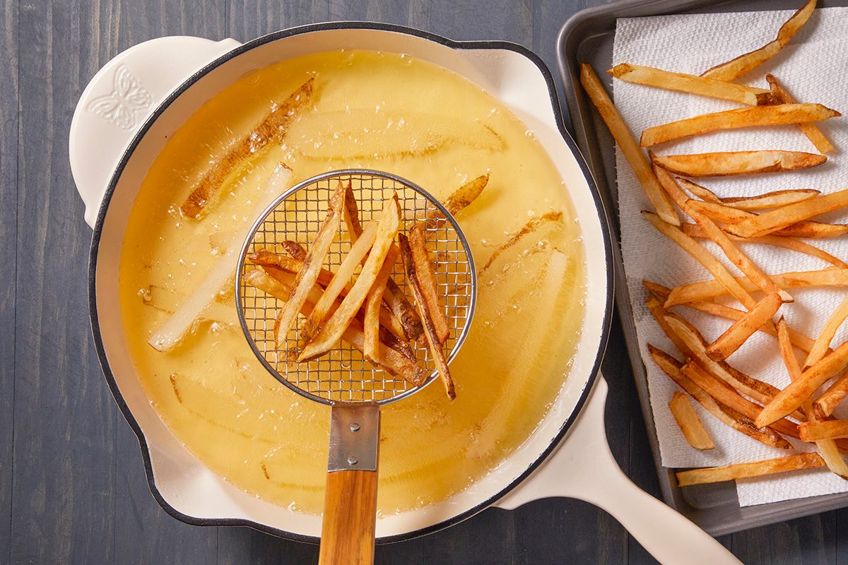 A white pan with hot oil is frying potato strips. A metal strainer lifts some fries out, while cooked fries drain on paper towels in a tray beside the pan.