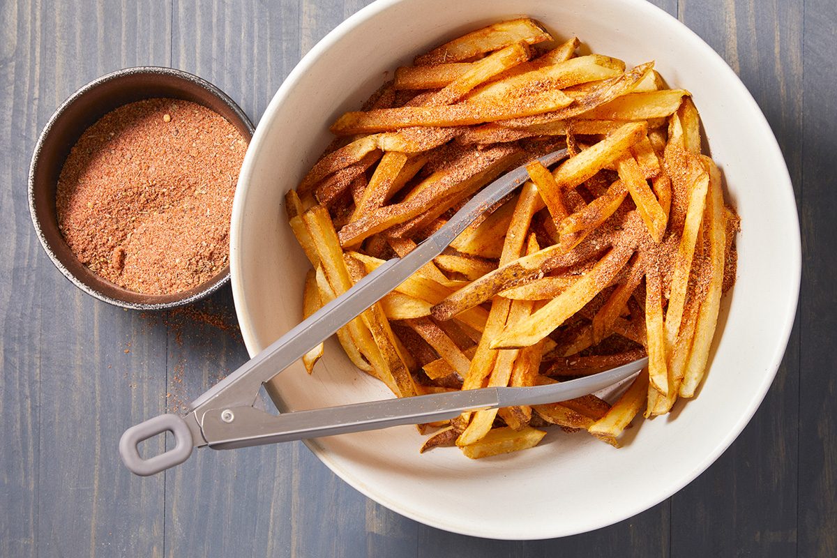 A white bowl filled with seasoned French fries and metal tongs, next to a small metal dish containing reddish-brown seasoning, set on a wooden surface.