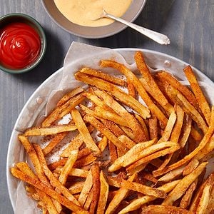 A plate of crispy seasoned French fries on parchment paper, served with a bowl of ketchup and a bowl of creamy dipping sauce with a spoon.