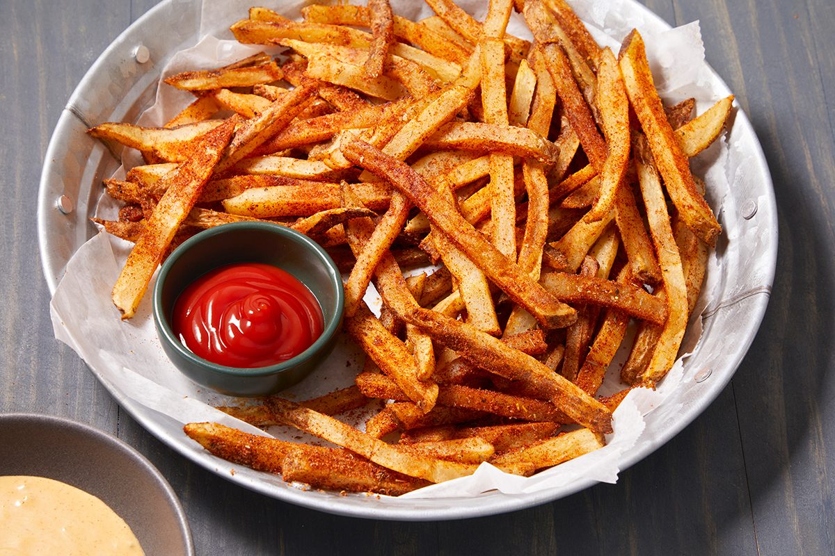 A plate of seasoned French fries served on parchment paper with a small bowl of ketchup on the side. Another bowl with a yellow dipping sauce is partially visible in the corner.