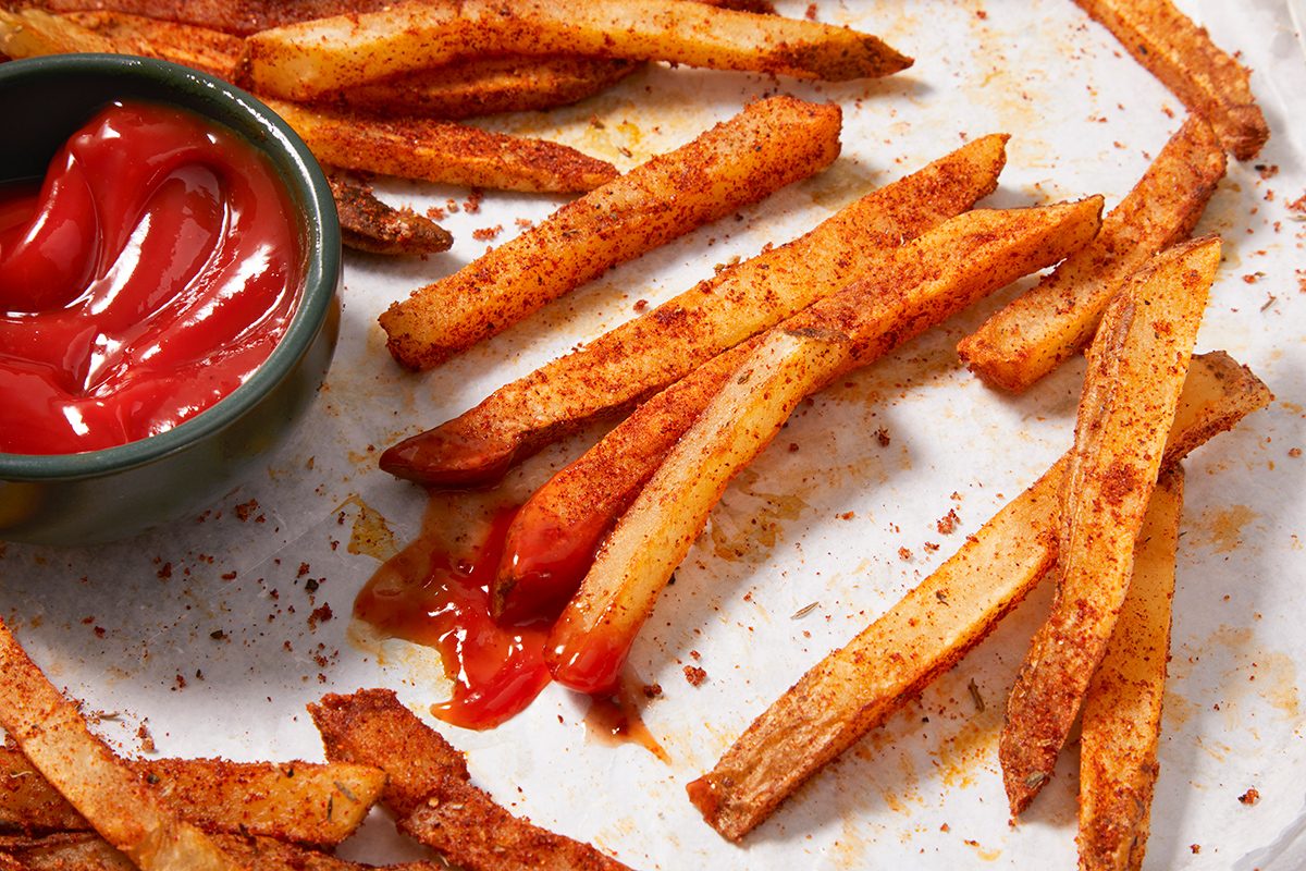 Seasoned French fries scattered on a parchment-lined surface with a small green bowl of ketchup. Some fries are dipped in ketchup, and a reddish seasoning is visible on the fries and paper.