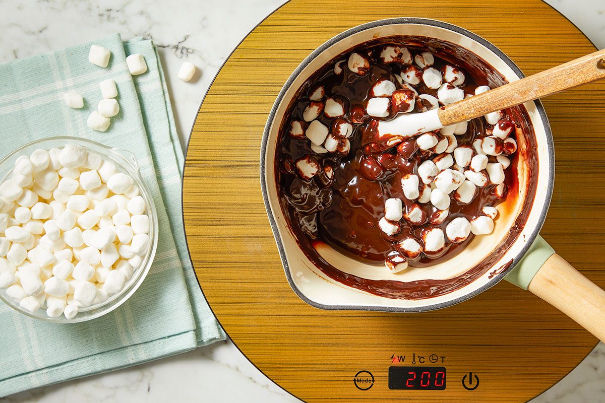 Overhead horizontal image of melted chocolate in a saucepan being mixed with marshmallows with a white spatula. A bowl of marshmallows sits to the side on a blue and white checkered napkin.