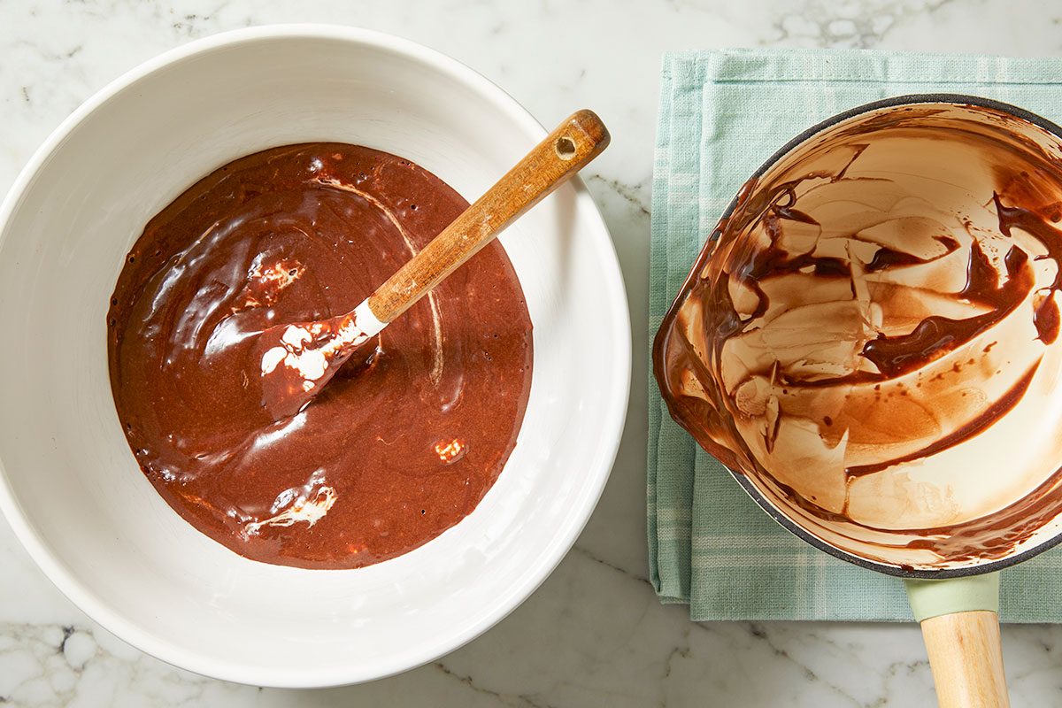 Overhead horizontal image of chocolate batter in a white bowl being mixed with a spatula. Next to the bowl is a saucepan with melted chocolate inside, sitting on a light blue napkin.