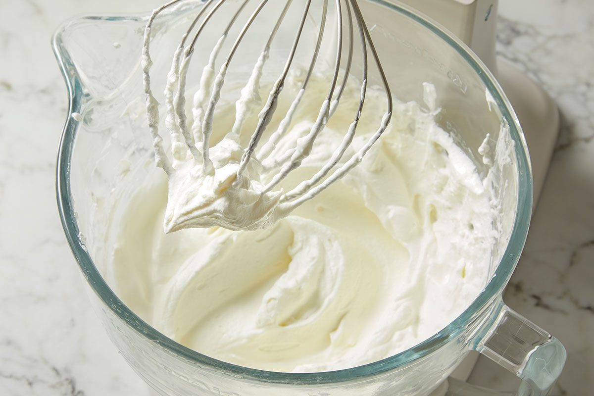 Overhead horizontal image of whipped cream being made in a clear glass mixing bowl, using a metal whisk. The cream is fluffy and white, and the bowl is sitting on a marble countertop.