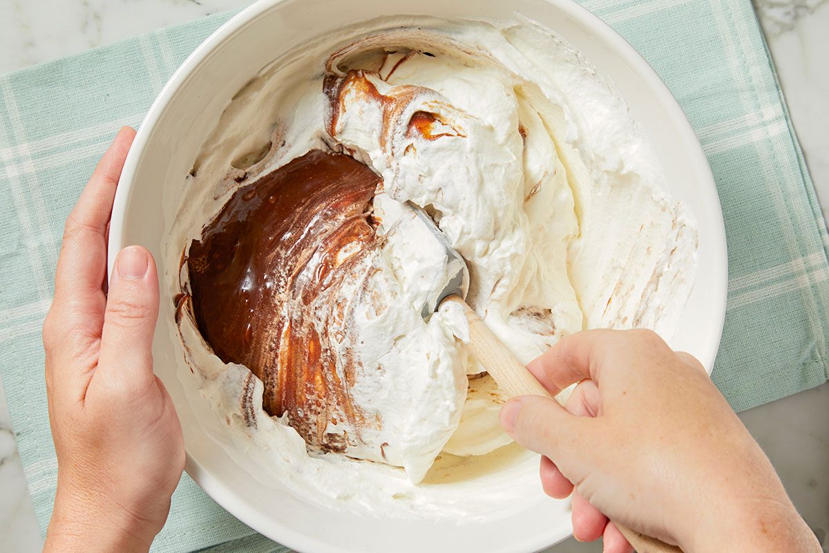 Eye Level horizontal image of a white bowl filled with whipped cream and chocolate being mixed with a spatula. Hands holding the bowl and spatula are visible.