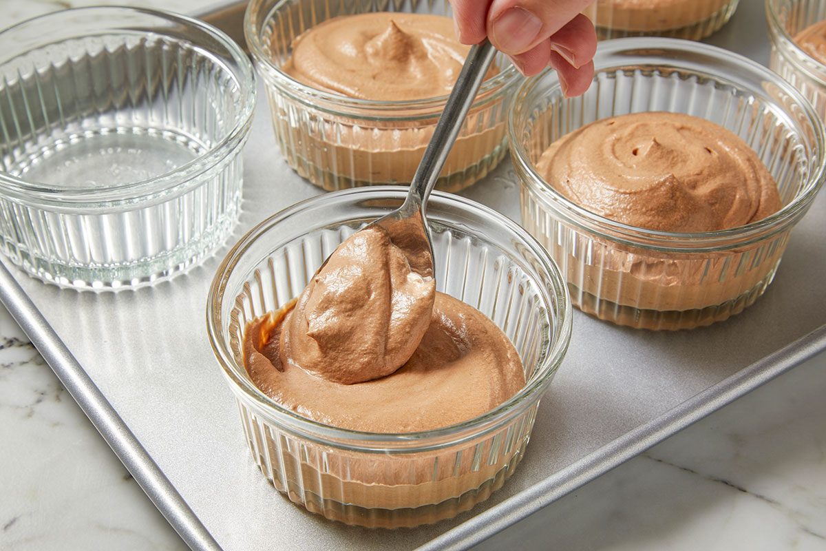 Eye Level horizontal image of a hand using a spoon to put chocolate mousse into a clear glass ramekin. Several ramekins, some filled with mousse, are sitting on a metal baking sheet.