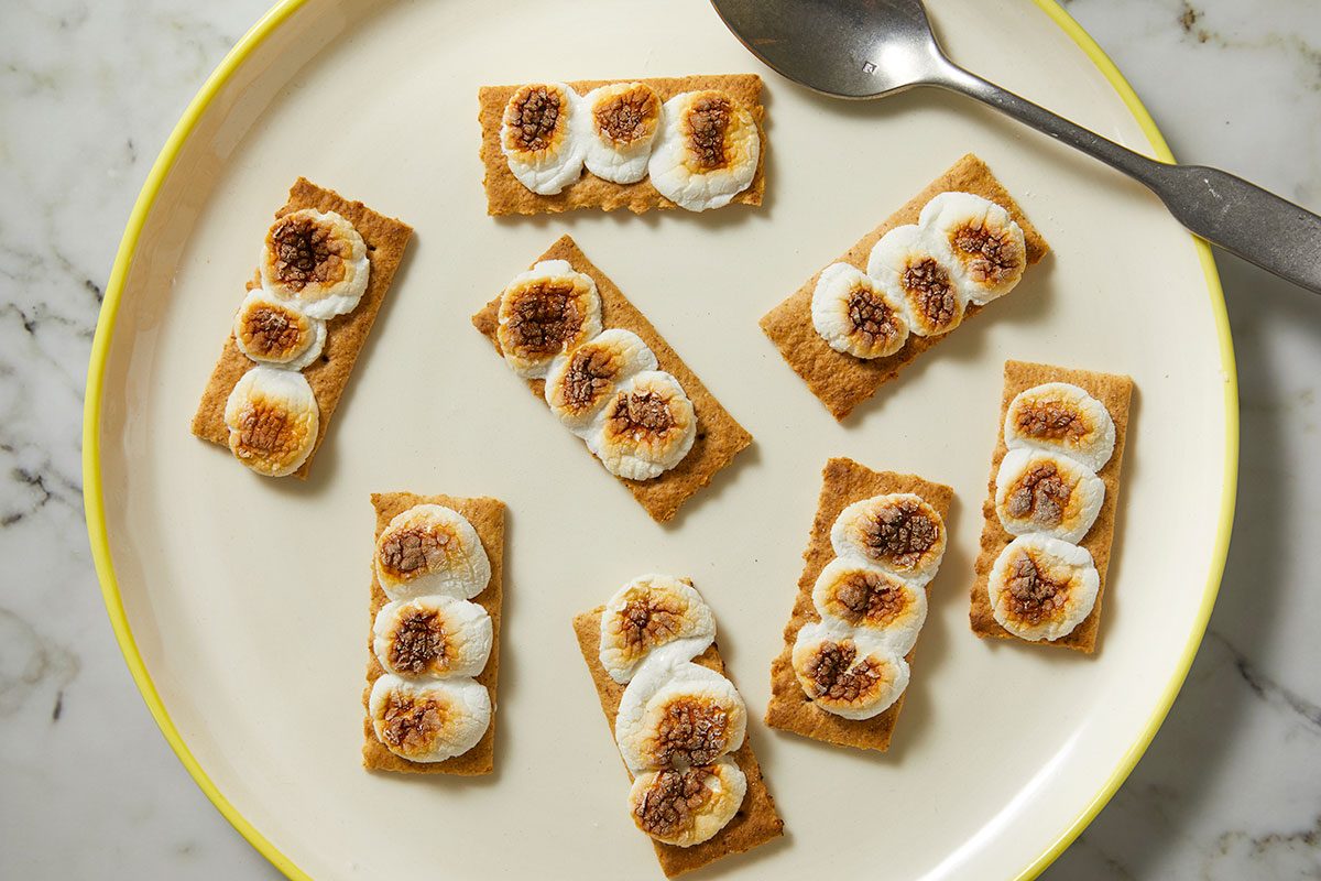 Overhead horizontal image of several graham crackers topped with toasted marshmallows, arranged on a yellow-rimmed plate, and a spoon is visible. The toasted marshmallows are browned and bubbly.