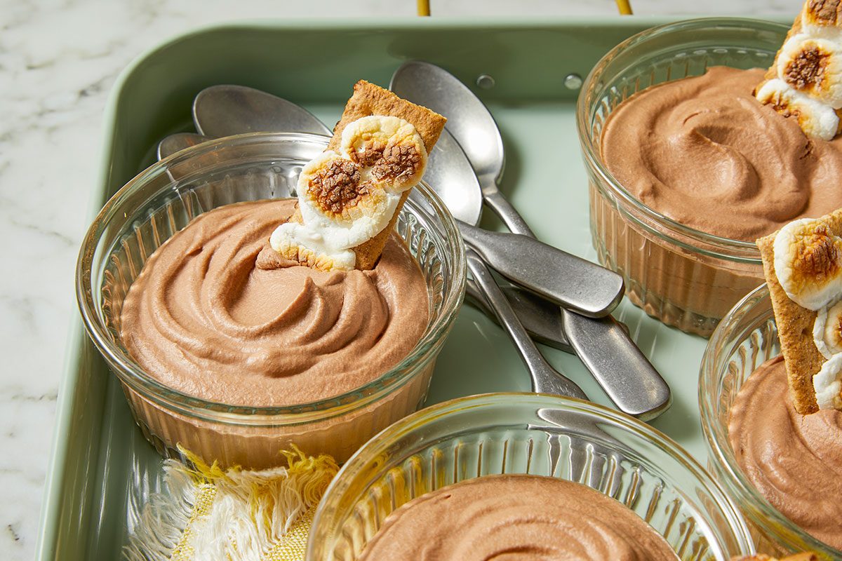 Eye Level horizontal image of S'mores Chocolate Pudding dessert in clear glass bowls with spoons, on a green tray. A toasted marshmallow on a graham cracker is stuck into the chocolate pudding.