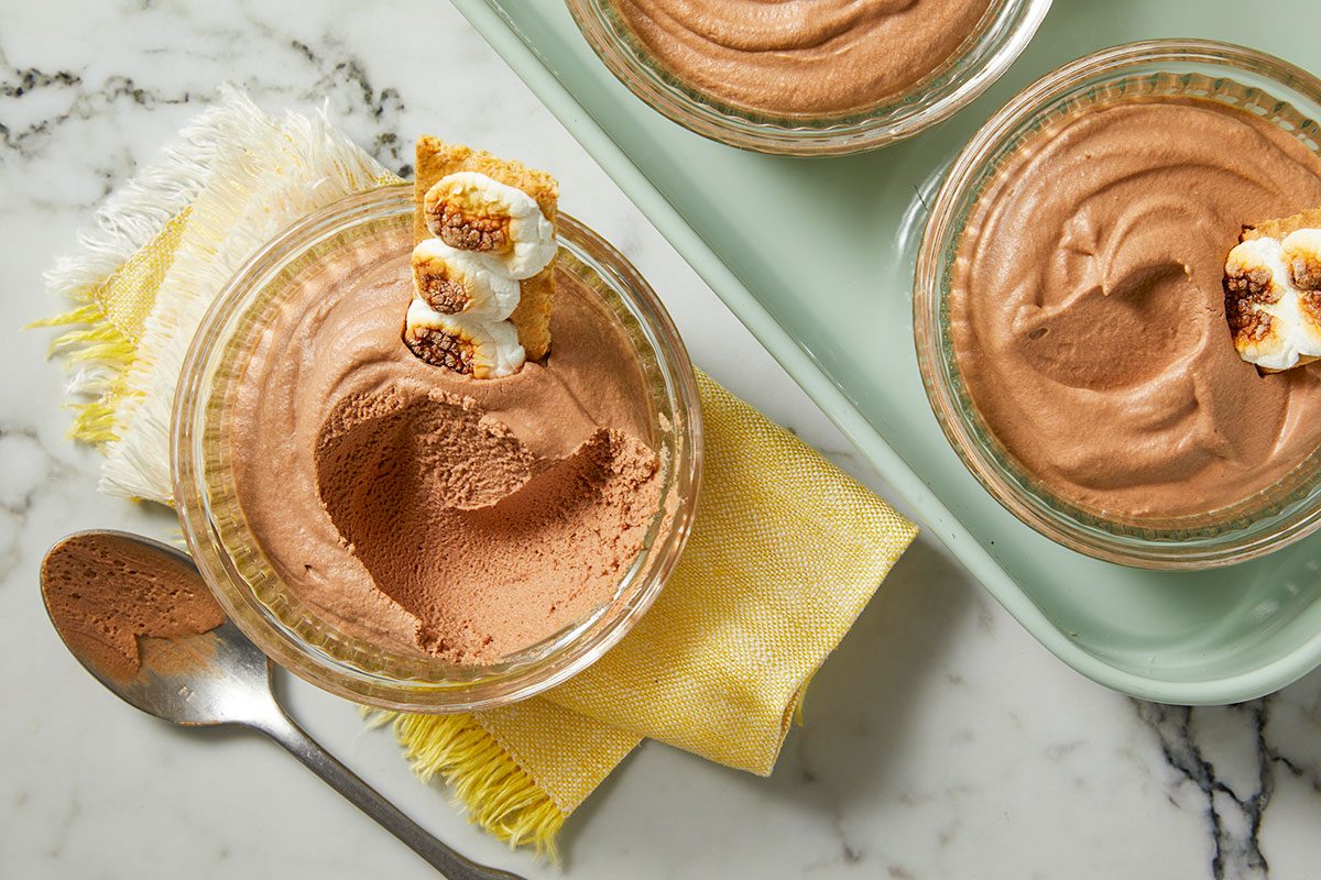 Overhead horizontal image of three s'mores chocolate mousse desserts in glass bowls. One dessert is garnished with toasted marshmallows and graham cracker and resting on a yellow and white fringed linen.