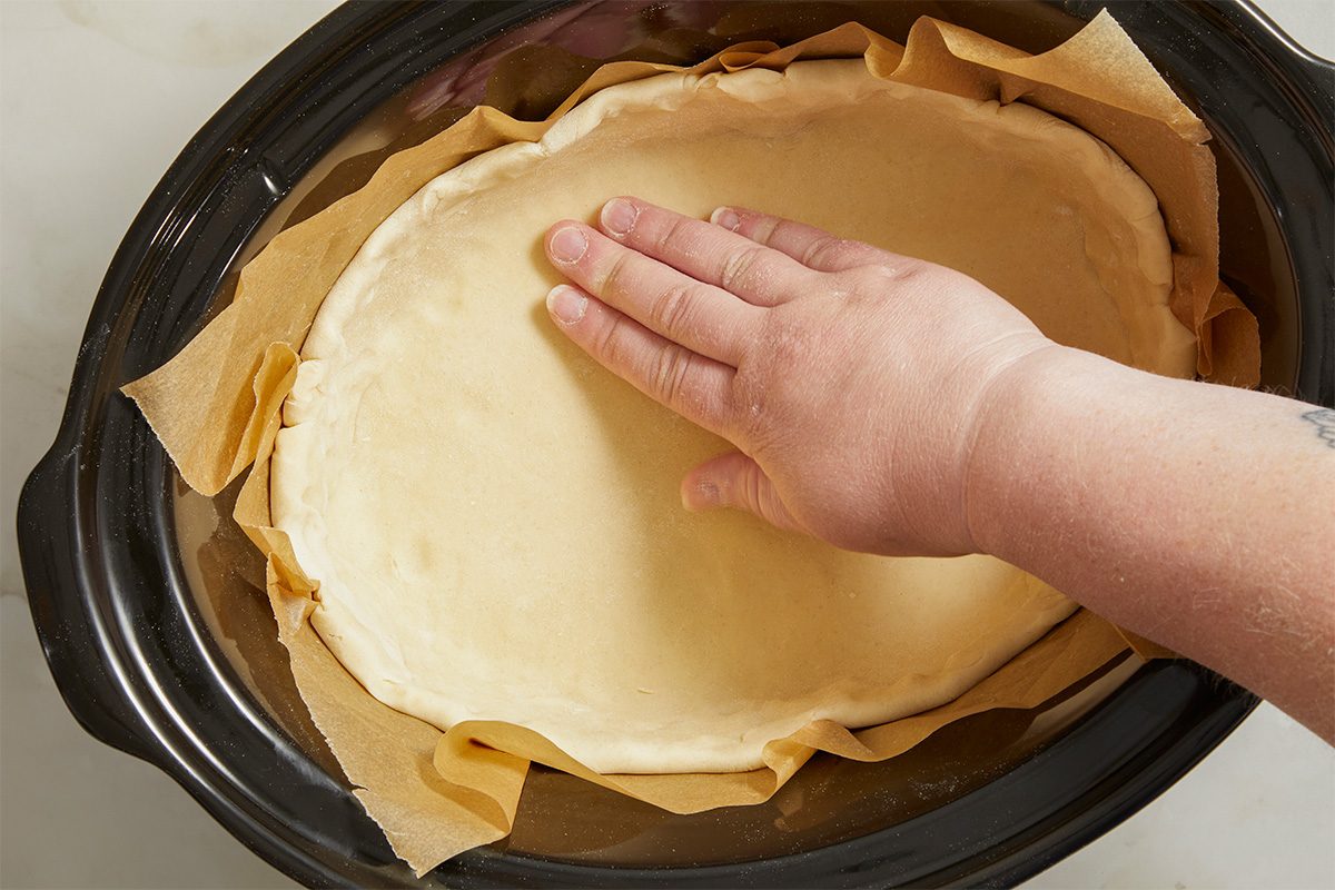 Overhead horizontal image of a sheet of refrigerated pie crust being pressed into a slow cooker lined with parchment paper. The baker's hand is pressing the pie crust to fit the bottom of the slow cooker.