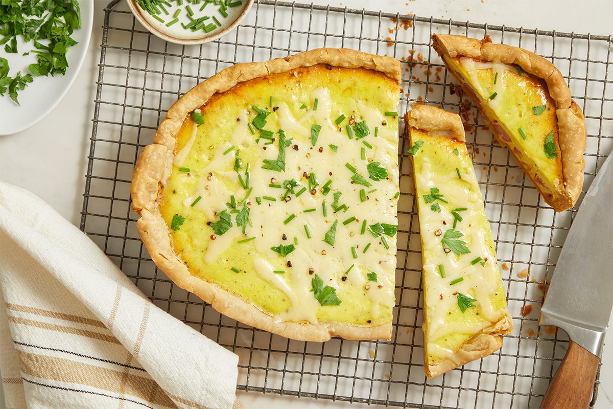 Overhead horizontal image of Slow-Cooker Ham & Gruyere Quiche. A partial quiche with two slices cut out is sitting on a wire rack, garnished with chopped chives and parsley. A knife is sitting next to one of the slices.