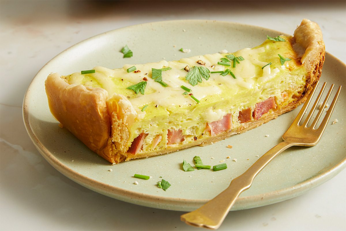Eye Level horizontal image of a slice of Slow-Cooker Ham & Gruyere Quiche on a green plate with a gold fork. Diced ham and shredded Gruyere cheese are visible in the slice of quiche, which is garnished with herbs.