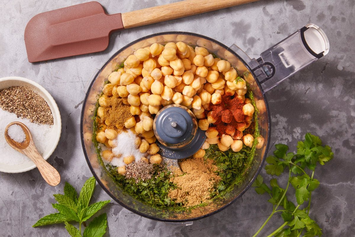 Overhead horizontal image of a food processor filled with chickpeas, herbs, spices and salt ready to be blended. Additional herbs and spices are to the side, with a spatula.