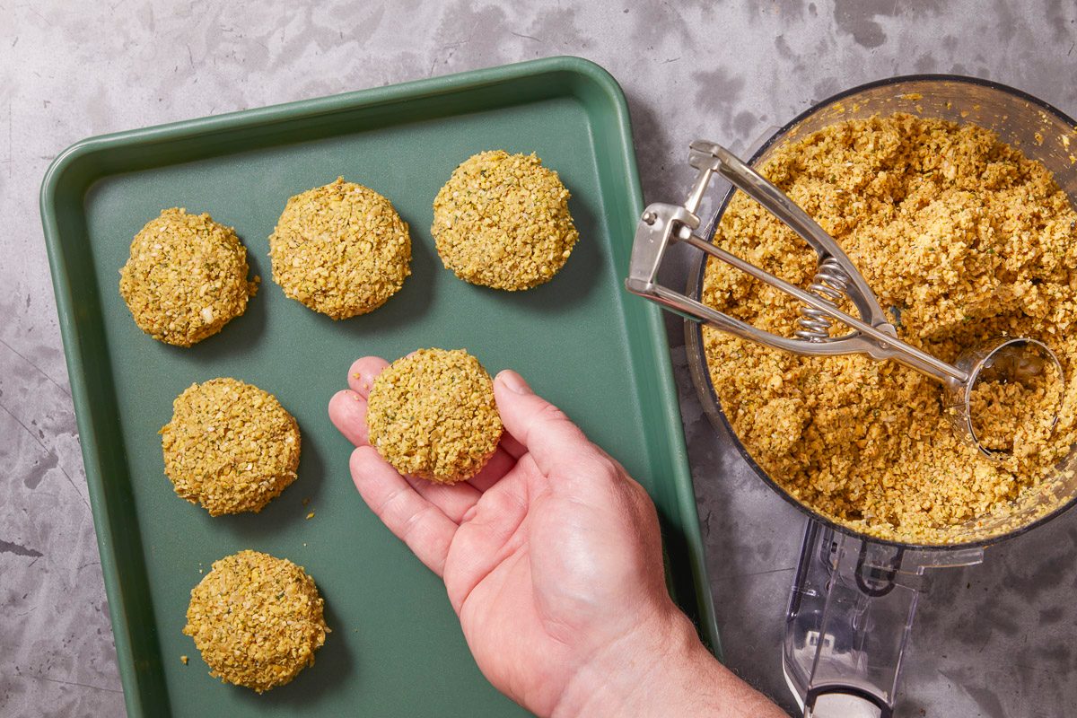 Overhead horizontal image of falafel being formed. A hand holds a formed falafel ball above a green baking sheet with five other falafel balls. Beside the sheet is a bowl of uncooked falafel mixture with an ice cream scoop in the mixture.