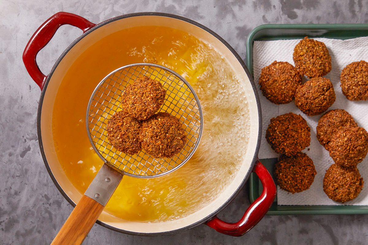 Overhead horizontal image of falafel being fried in hot oil using a wire skimmer in a red cast iron pot. More fried falafel are seen resting on a metal tray lined with white paper towels to drain excess oil.