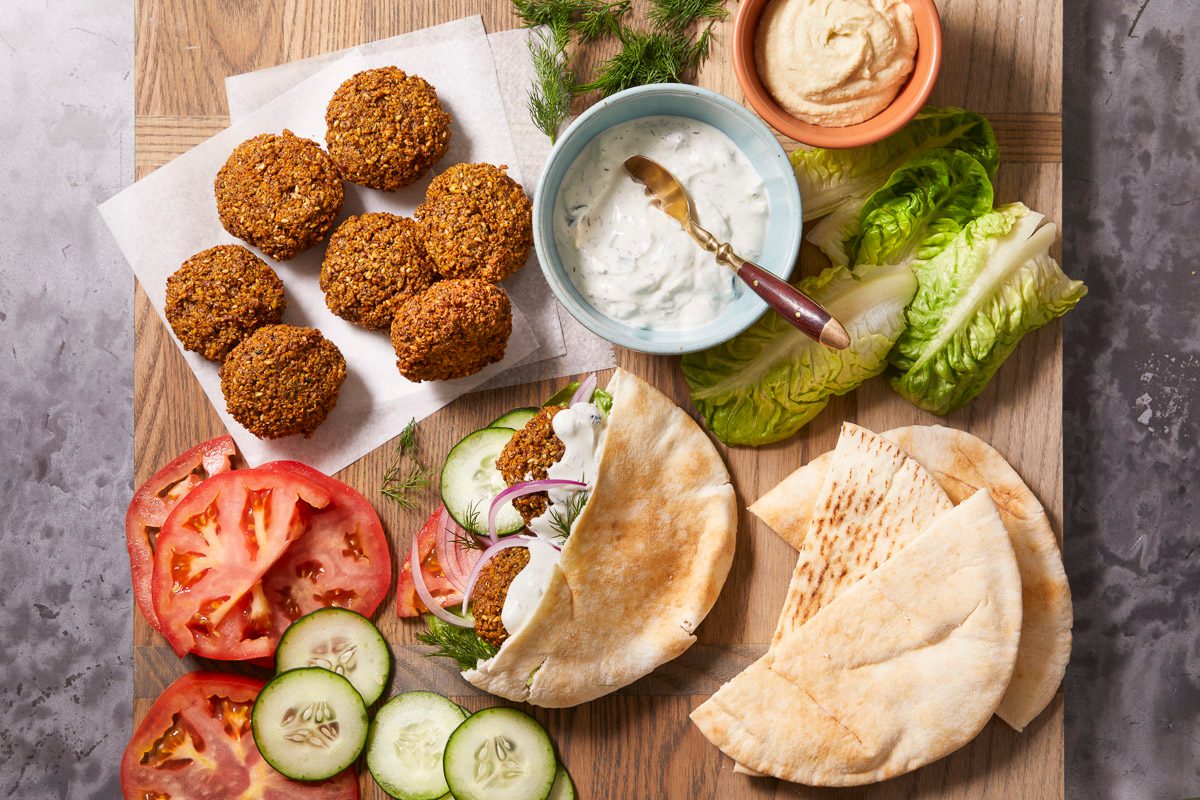 Overhead horizontal image of falafel on a wooden cutting board. Falafel are arranged on parchment paper, and a pita is stuffed with more falafel, cucumbers, red onion, lettuce, and white sauce. Bowls of white sauce and hummus sit nearby.