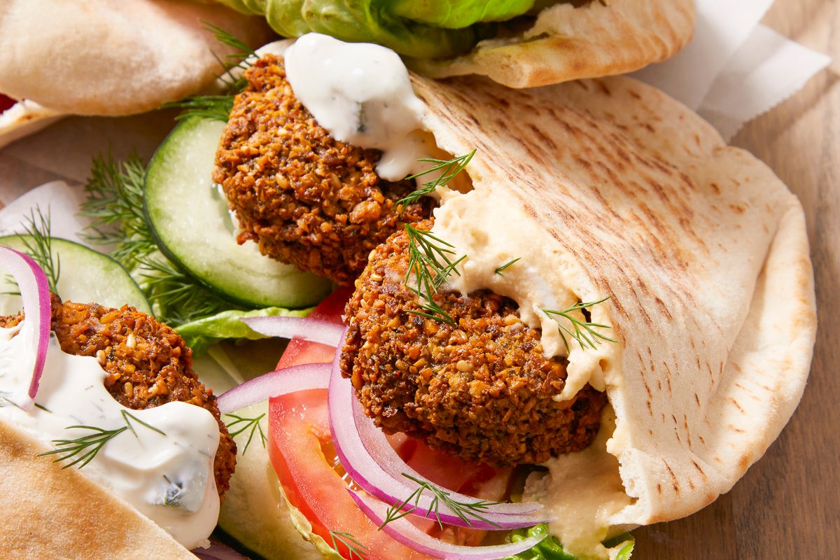 Eye Level horizontal image of a falafel pita sandwich. The sandwich is filled with falafel, lettuce, sliced red onions and cucumbers, tomatoes, and topped with a white sauce and dill.