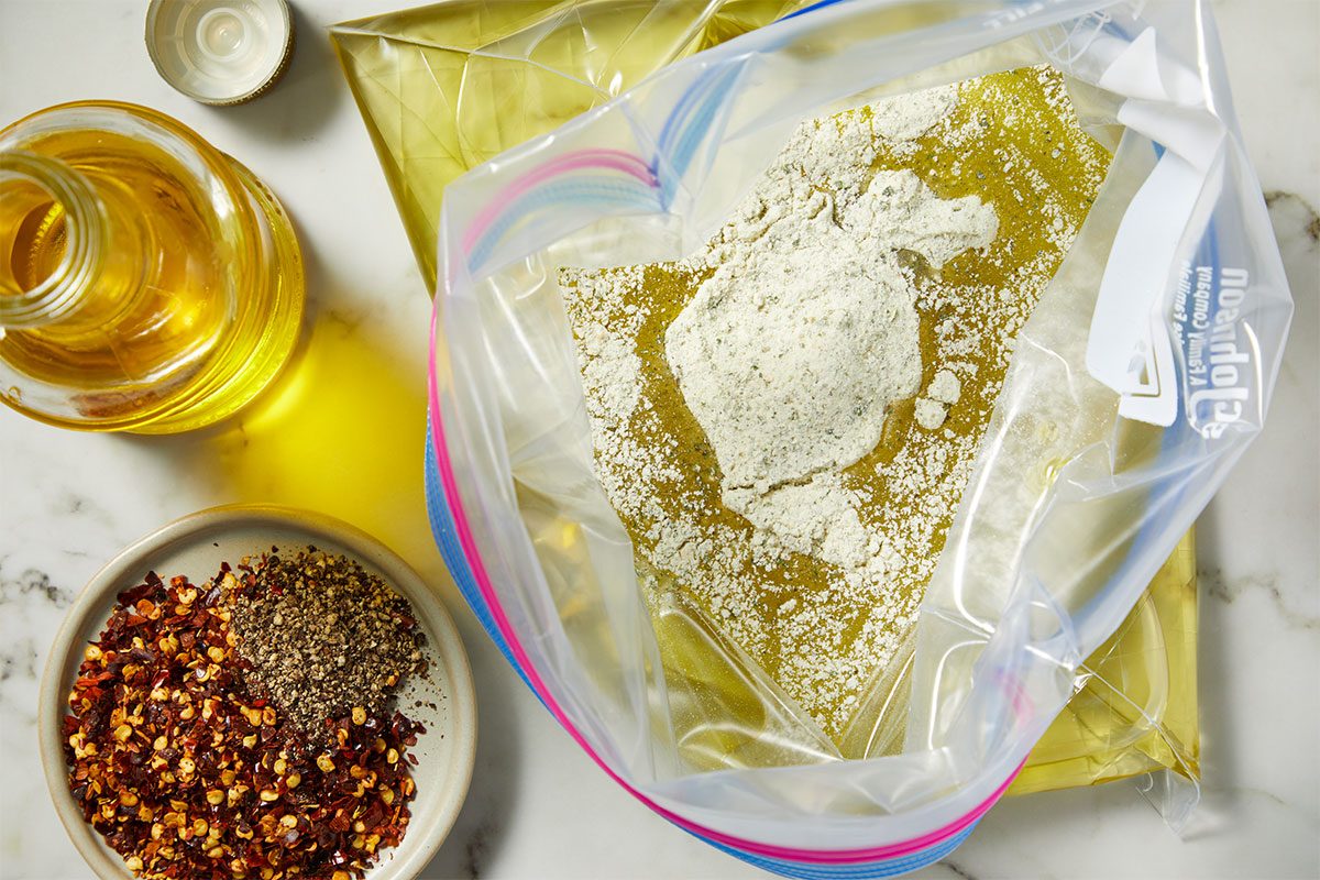 Overhead horizontal image of a Ziploc bag open to reveal a liquid base covered with a white powdered seasoning mixture. Adjacent to the bag are an uncapped bottle of oil and a small dish containing red pepper flakes and black pepper.