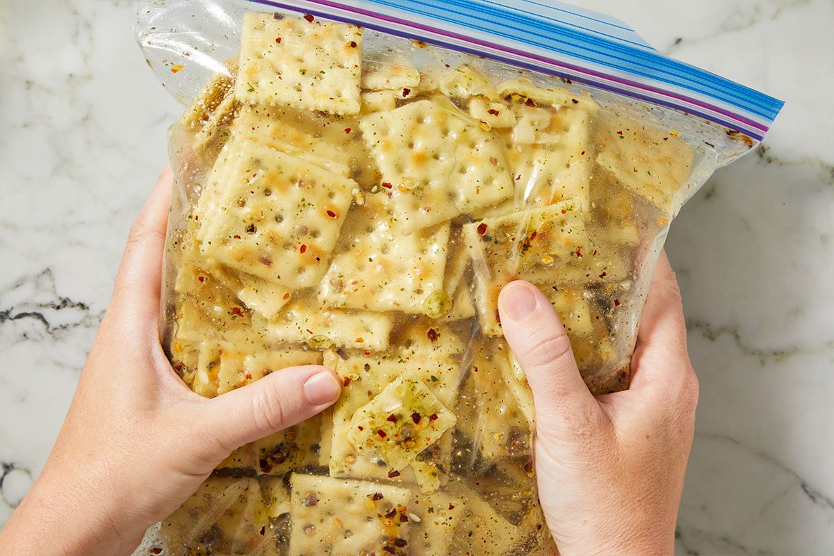 Overhead horizontal image of two hands holding a clear plastic bag filled with square crackers. The crackers are thoroughly coated in a vibrant, seasoned oil mixture, featuring visible green herbs and red pepper flakes. The bag is being held against a white marble countertop.