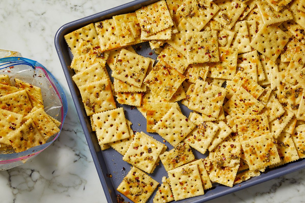 Overhead horizontal image of seasoned crackers in a baking pan. The square crackers are coated with an oily mixture, speckled with red pepper flakes and green herbs, and piled high in a dark gray baking sheet. A plastic bag of similar crackers sits to the left on a marble countertop.