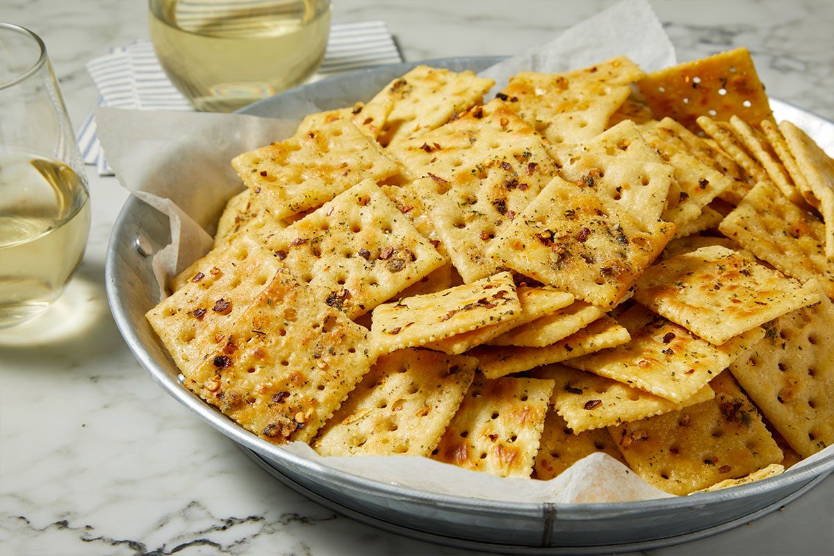 Eye Level horizontal image of a metal tin filled with golden-brown seasoned crackers. The crackers are sprinkled with red pepper flakes and green herbs, sitting on parchment paper within the tin. Two glasses of white wine are visible in the soft-focus background.