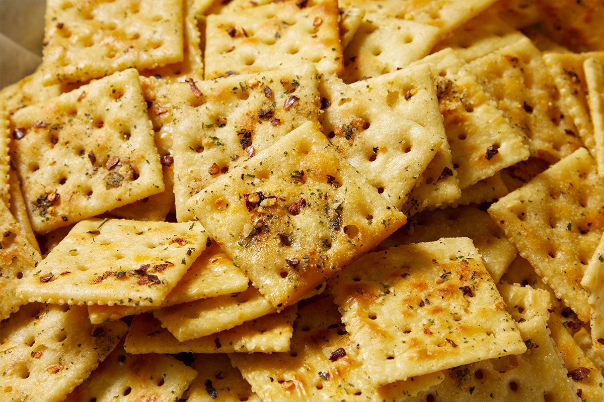 Overhead horizontal image of a large pile of seasoned crackers. The golden-brown crackers are heavily coated with green herbs and red pepper flakes, showcasing a savory snack.