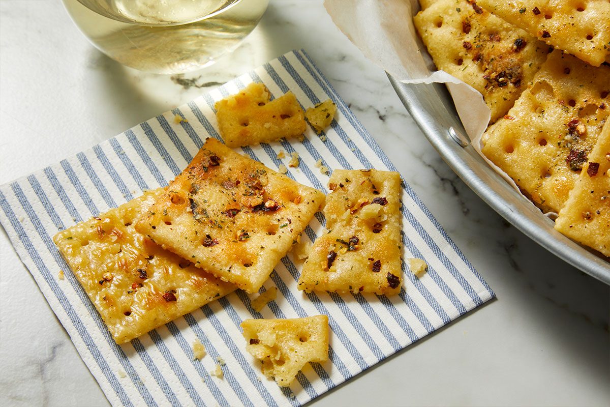 Overhead horizontal image of seasoned crackers on a blue and white striped napkin, with more crackers visible in a metal serving basket lined with parchment paper. The crackers are sprinkled with herbs and red pepper flakes, and a glass of white wine is in the background.