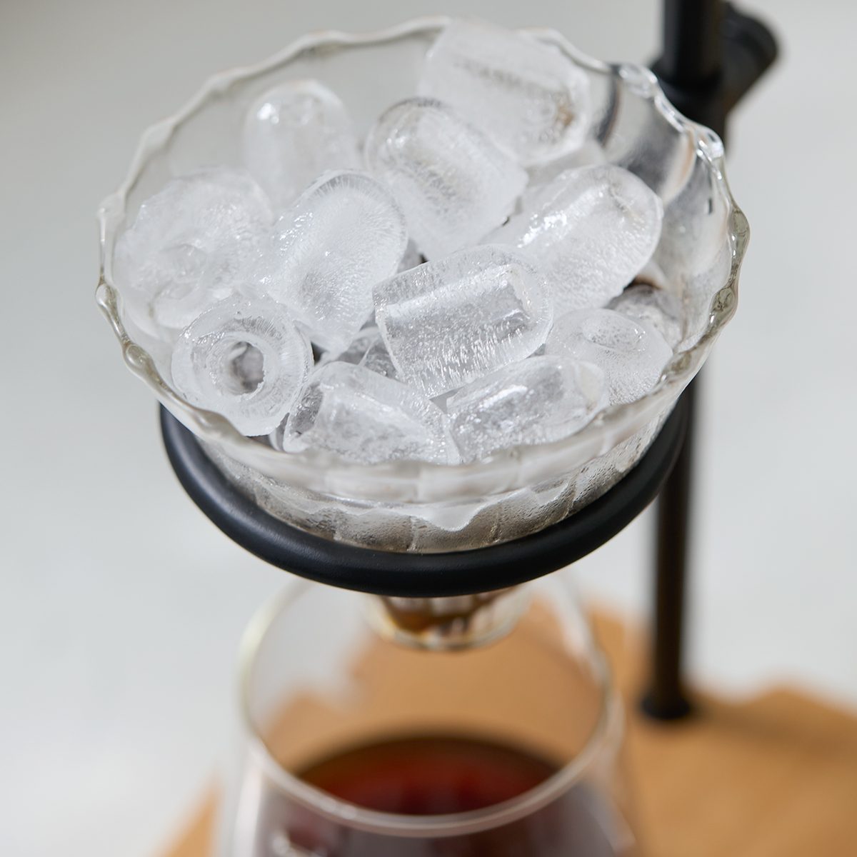 A glass bowl filled with ice cubes sits above a glass carafe containing a dark liquid, likely coffee, in a cold brew drip coffee maker setup.