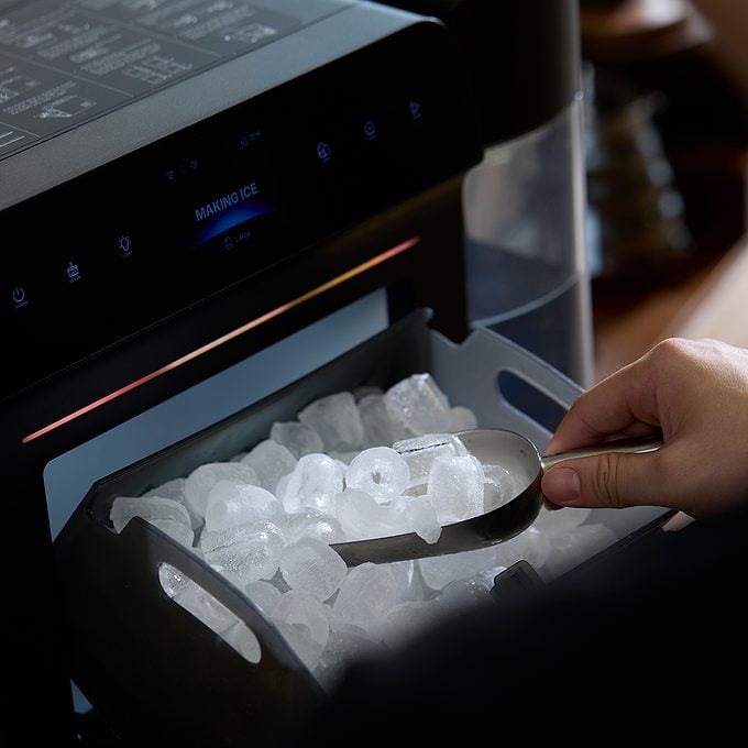 A hand uses a metal scoop to collect ice cubes from the ice maker compartment of a modern refrigerator.