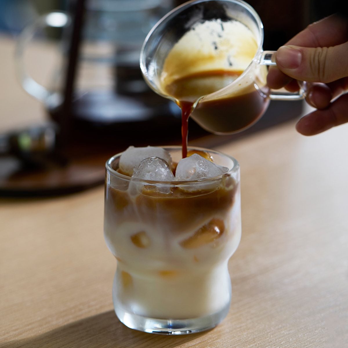 A hand pours espresso from a small glass pitcher over a glass filled with ice and milk, creating an iced latte on a wooden table.