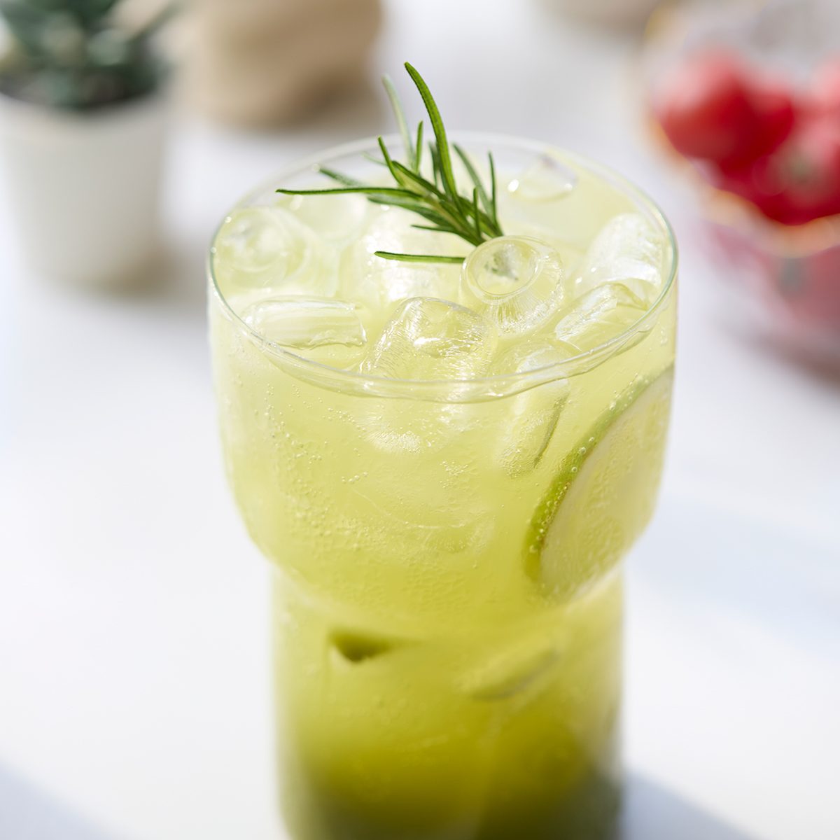 A glass filled with a green, iced beverage garnished with a sprig of rosemary and a lime slice, set on a bright surface with blurred objects in the background.