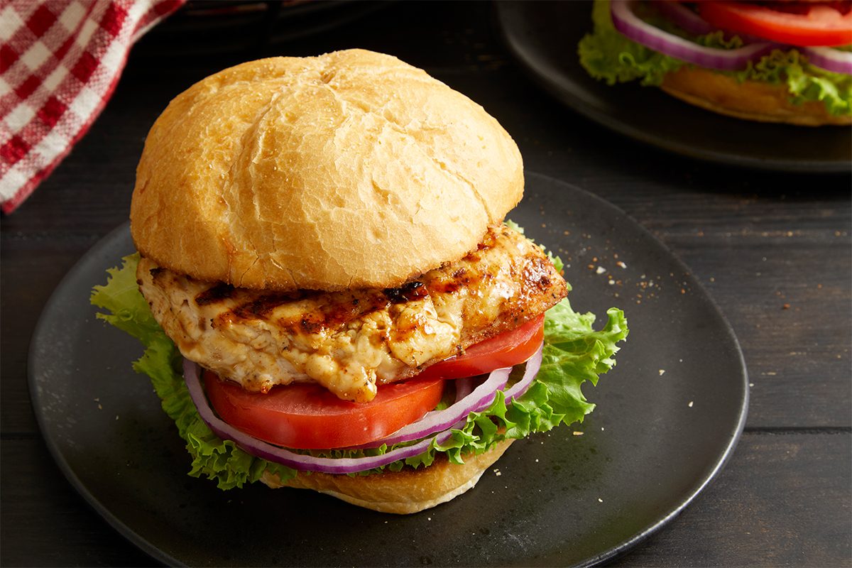 Eye Level horizontal image of a fully assembled Cajun Chicken Sandwich on a black plate. The sandwich features a grilled chicken breast, sliced tomatoes, red onions, and lettuce leaves between two halves of a kaiser roll. Another sandwich is visible blurred in the background.