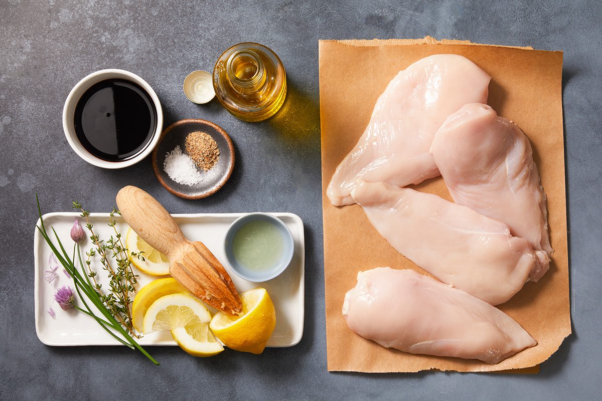 Raw chicken breasts on parchment paper next to small bowls of soy sauce, oil, spices, fresh ginger, lemon slices, garlic, herbs, and lime juice, all arranged on a gray surface.