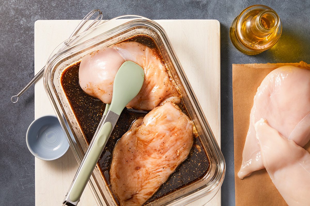 Raw chicken breasts marinating in a glass dish with marinade, a green-tipped spatula resting on top. Nearby are more raw chicken breasts on paper, a small bowl, a whisk, and a bottle of oil.