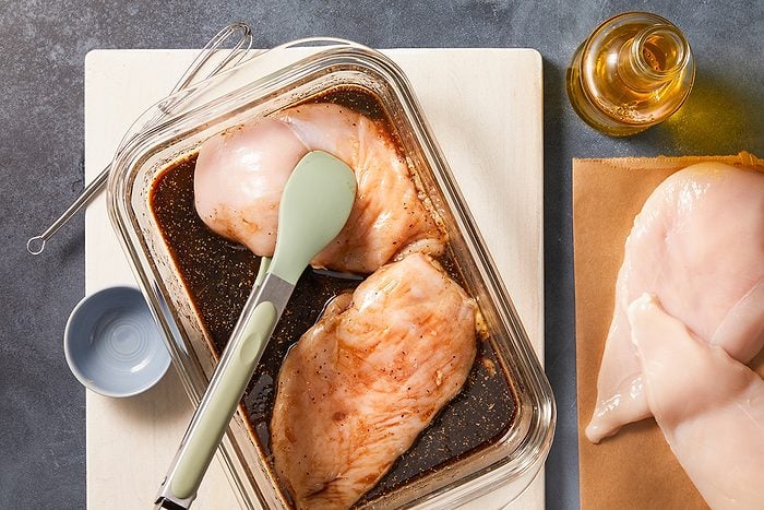 Raw chicken breasts marinating in a glass dish with marinade, a green-tipped spatula resting on top. Nearby are more raw chicken breasts on paper, a small bowl, a whisk, and a bottle of oil.