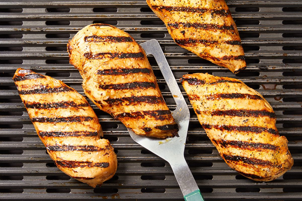 Four grilled chicken breasts with visible char marks are cooking on a grill. A metal spatula is placed under one of the chicken breasts, ready to flip or serve it.