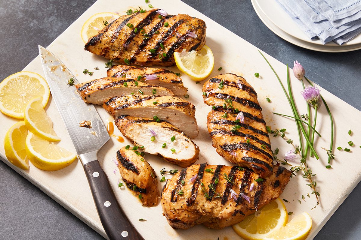 Grilled chicken breasts sliced on a cutting board, garnished with fresh herbs and lemon slices. A knife rests beside the chicken, and a plate with napkins is visible in the background.