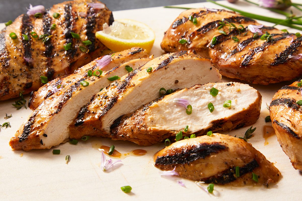 Grilled chicken breasts with char marks, sliced and garnished with chopped herbs, are arranged on a light-colored cutting board with a lemon wedge in the background.