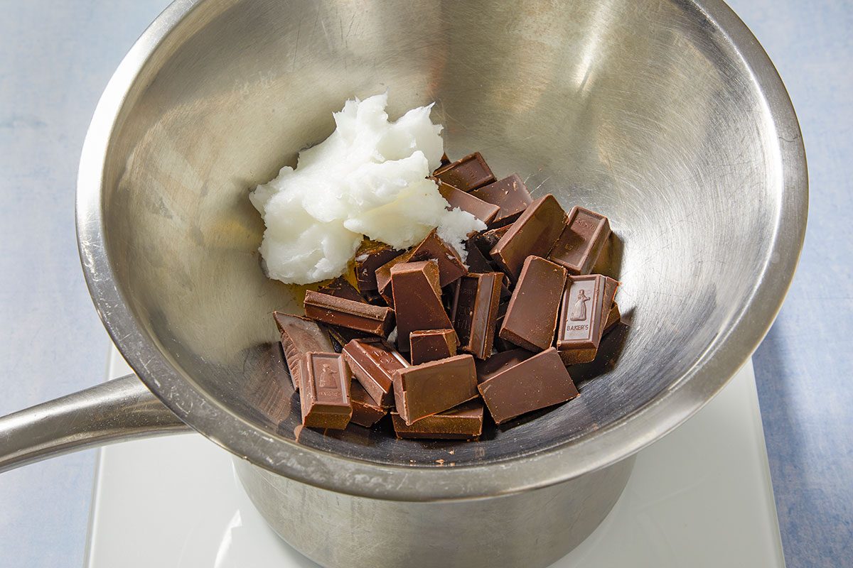 Overhead horizontal image of chunks of Baker's dark chocolate and a scoop of white coconut oil or fat in a stainless steel bowl. The bowl is placed over another pot, likely for melting the ingredients.