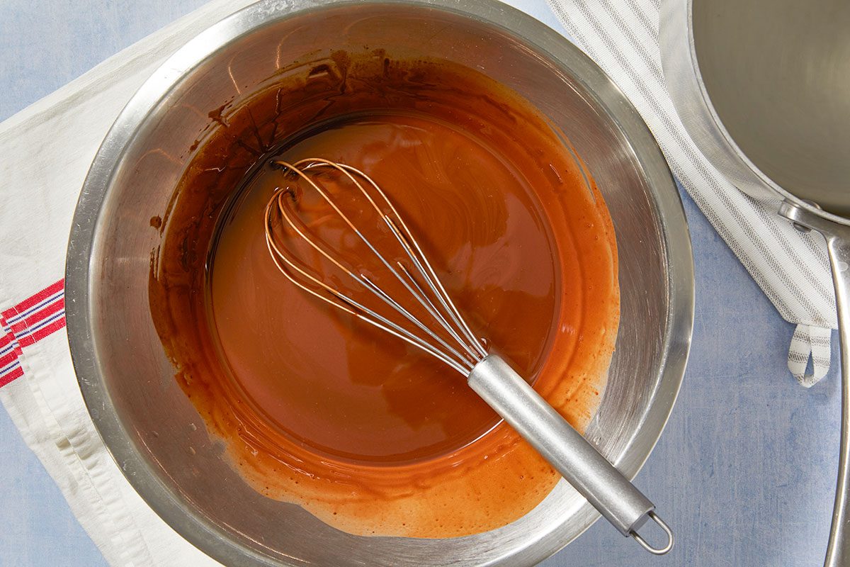 Overhead horizontal image of a stainless steel mixing bowl filled with a rich, dark brown liquid, resembling caramel or chocolate sauce. A metal whisk rests within the viscous mixture, suggesting preparation. A white kitchen towel and part of a saucepan are visible around the bowl, all set on a light blue surface.