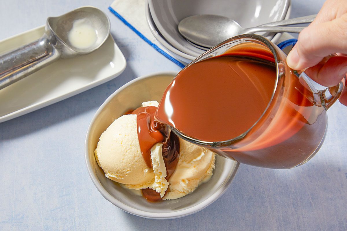 Eye level horizontal image of a hand pouring rich chocolate sauce over two scoops of vanilla ice cream in a gray bowl. An ice cream scoop with a bit of melted ice cream rests on a white dish in the background, next to stacked bowls and spoons.