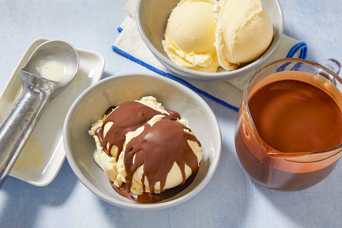 Overhead horizontal image of two bowls of vanilla ice cream on a light blue surface. One bowl of ice cream is topped with chocolate hard shell, while the other remains plain. A pitcher of chocolate sauce and an ice cream scoop are also visible.