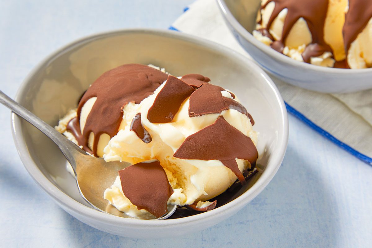 Eye Level horizontal image of a bowl of vanilla ice cream topped with a chocolate hard shell, with some pieces flaking off. A metal spoon is resting in the bowl, with another blurred bowl of ice cream in the background on a blue and white patterned napkin.
